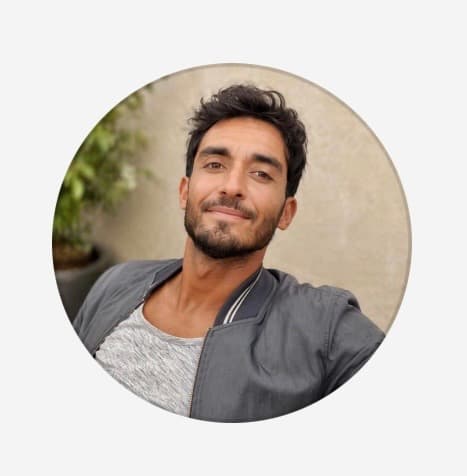 Smiling man with dark wavy hair and a beard, wearing a grey bomber jacket.